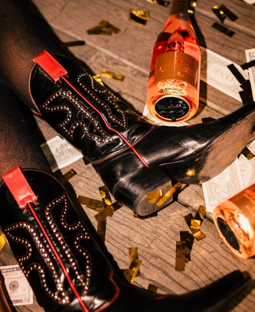 Two legs wearing cowboy boots and a bottle of bubbly on the floor of a party